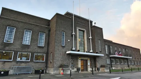 Local Democracy Reporting Service A landscape shot of Havering Town Hall. It is a wide, dark brown brick building with long windows, in an international modern architectural style.