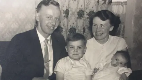 William Cruickshank A black and white picture of William Cruickshank, centre, as a child with his family. His father, William Crocket is on the left of the picture, wearing a dark suit, light shirt and striped tie. William is in the middle and is wearing a light T-shirt shirt. His mother, wearing a light blouse is on the right, holding William's sister, who is a baby and wearing a light top. They are sitting in front of curtains with a floral pattern.