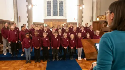 North Yorkshire Council Children wearing purple jumpers sing in a church, led by a woman stood at a lectern.