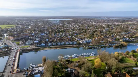 Getty Images An aerial view of East Molesey which shows the River Thames in the foreground and boats and houses along the edge of the river. Hampton Court bridge is seen in the left of the image and in the distance there are reservoirs and blue sky.