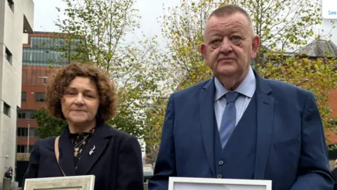 A woman and a man pictured outside a building. The woman is on the left, she is middle-aged with short brown curly hair and is wearing a blue jacket. The man on the right is older, with a blue suit, blue tie and blue shirt.