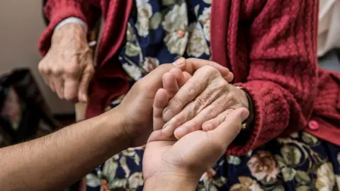 Getty Images A pair of hands holding an elderly woman's hands. The woman wears a red cardigan and floral dress. She is seated.
