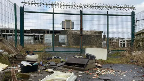 BBC A discoloured fridge door lying on its back and a broken brown, wooden bit of school furniture are among the detritus dumped outside the green metal gates of the former school. 