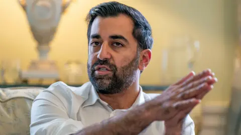 PA Media Humza Yousaf sitting in a chair in a yellow-lit room. He is wearing an open-necked white shirt and is clearly speaking and using his hadns to make a point. He is holding them together.