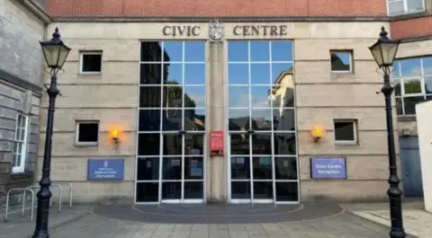 BBC The Civic Centre in Stoke, a light-coloured brick building with large panelled windows which are about two-storeys in height. There are two traditional-style lampposts in the foreground.
