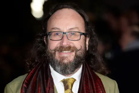 Getty Images Dave Myers, photographed arriving at the National Television Awards 2015. He is balding but has long hair at the back, square glasses, a greying beard and a waxed moustache. He is wearing a greenish jacket, a white shirt and a spotted tie to match the jacket. He also is wearing a dark red silk scarf.