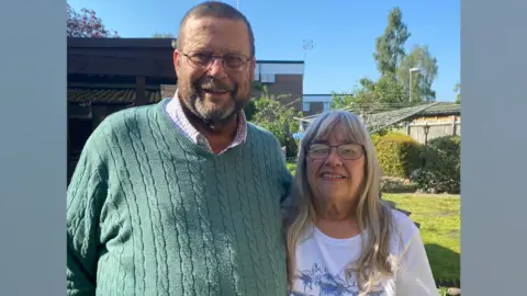 Family photo Kevin and Sue Starling. Kevin, on the left, appears in a checked shirt and green jumper over the top. His wife also wears glasses, and has a white t-shirt on. They are standing in a garden