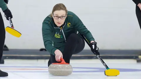 World Curling/Anil Mungal A woman in a green Australia top holding a curling broom prepares to send a red curling stone down the ice