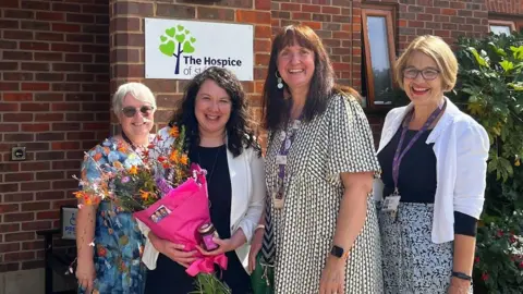 Victoria Collins Three ladies in summer outfits pose with MP Victoria Collins outside the entrance of the hospice. Ms Collins is holding a bunch of flowers and a jar of preserve.
