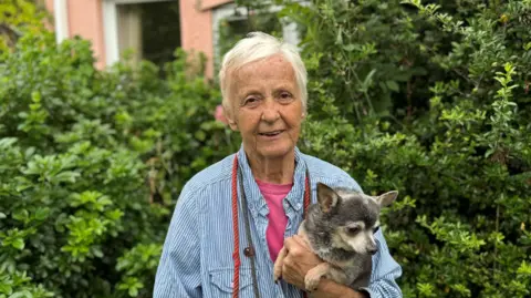 George King/BBC Maggie Jackaman, who has short grey hair and is wearing a light blue and white striped shirt over a pink top, holding her dog, Kenny. She is standing in front of a hedge.