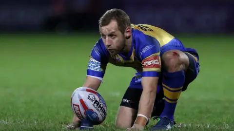 PA Media Rob Burrow in his blue and yellow Leeds Rhinos kit as he prepares to kick a ball on a green pitch