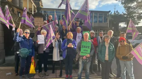 George Torr/BBC People outside County Hall in Matlock protesting against the closures. Some are waving purple UNISON flags.