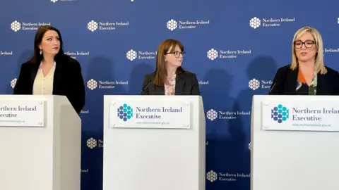 PA Deputy First Minister Emma Little-Pengelly, Finance Minister Caoimhe Archibald and First Minister Michelle O'Neill in black blazers, stood in front of Northern Ireland Executive white podiums with microphones, against a blue backdrop