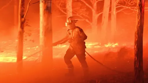 AFP A NSW RFS volunteer firefighter wearing full protective gear points a hose as he's surrounded by fire