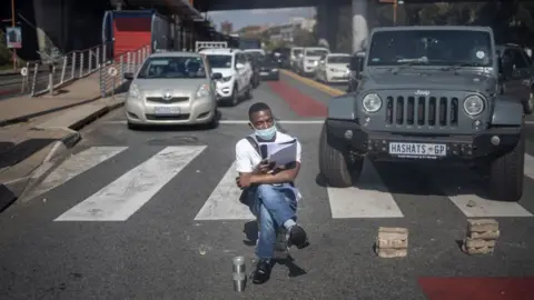 AFP A man sits on bricks pretending to read as students members of the African National Congress (ANC) and the Economic Freedom Fighters (EFF) block traffic in Braamfontein, Johannesburg, South Africa - Wednesday 10 March 2021