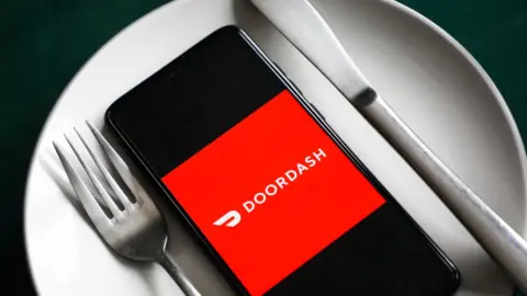 Getty Images A mobile phone showing the DoorDash logo sits between a fork and a knife on top of a dinner plate