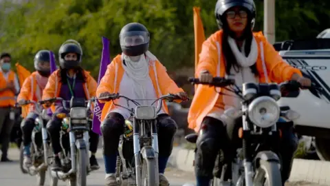 Getty Images Women in Pakistan ride motorbikes and wear helmets and orange jackets.