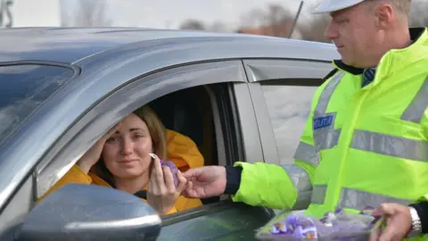 Romanian National Police Policeman hands purple flowers to woman through car window