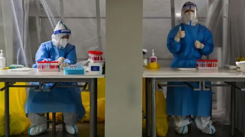 Getty Images Health workers wearing personal protective equipment wait to take swab samples from people during a Covid-19 coronavirus lockdown in the Xuhui district of Shanghai.