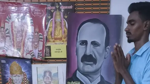Gubendran 20-year-old student offering prayers in front of images of Hindu gods and the colonial engineer John Pennycuick