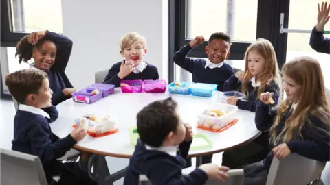 Getty/monkeybusinessimages Elevated view of primary school kids sitting together at a round table eating their packed lunches