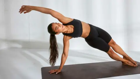 Rebecca Spencer Stef is wearing a black two piece sports bra and cycling shorts, her hair is tied back in a ponytail. She's holding a side plank post on a black exercise mat. Her eyes are closed and she's pouting her lips.