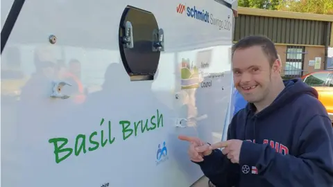 Teamwork Trust Side of a street sweeper bearing the legend "Basil Brush". A young man in a blue top is pointing to the name.