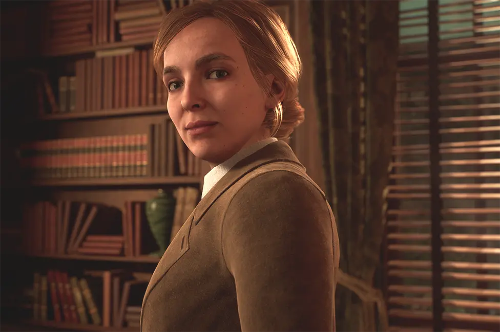 THQ Nordic A blonde woman with her hair tied back in a neat bun wears a smart tan jacket over an off-white blouse in this computer-generated portrait. She's looking at the viewer, smiling slightly. She's in an old-fashioned looking study or library - behind her are shelves stocked with volumes of books, all with matching spines and neatly arranged to signal where one collection ends and another begins.