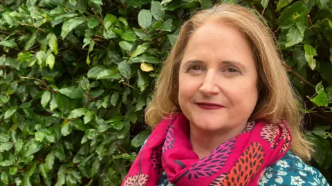 Amy Holmes/BBC Lara Pringle looks directly at the camera. She has light brown hair and is wearing a pink scarf with images of trees on it, and a green blouse with flowers on it. Behind her is a hedge.