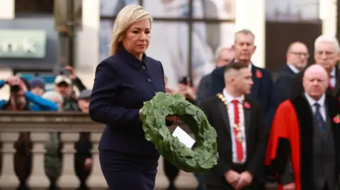 PA Media Michell O'Neill wearing a blue jacket carrying a green wreath. Behind her some men stand out of focus wearing suits and poppies. To her left behind stone columns some people hold up cameras and phone to take photos.