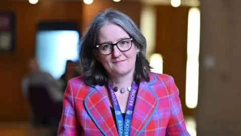 Getty Images Maree Todd wearing a red and purple tartan jacket and large-rimmed glasses walking through the Scottish Parliament building