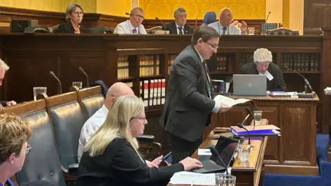 Lawrie Hooper standing up in the Tynwald chamber reading a speech. The chamber has wooden panels and book cases, and the Legislative Council can bee seen seated on the level above the MHKs.