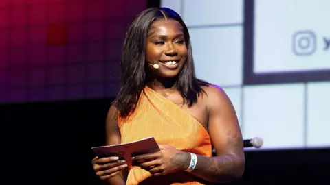 Tim Whitby Jade Barnett in an orange dress standing on stage. She holds notecards and wears a microphone. She is at the Youth Music Awards 2024.