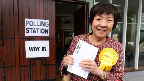Pacemaker Archive photo of Anna Lo pictured outside a polling station.  She is wearing a multicoloured jacket and a yellow Alliance rosette.  She is smiling and holding a polling card.