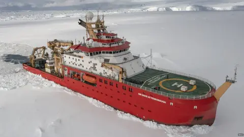 BAS The RSS Sir David Attenborough is a large red ship with a green helipad on the front. It is shown sailing through icy, snow-covered water