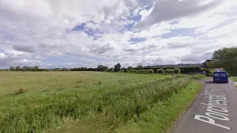 Google StreetView image of the land earmarked for development. There is a single-track road on the right of the picture, and to the left an open green field with power lines running over it. 