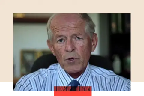 Getty Images A close-up of John Smyth speaking directly to the camera. His mouth is slightly open, and he is dressed in a white and blue striped shirt. He sits on a black chair, with a blurred background.