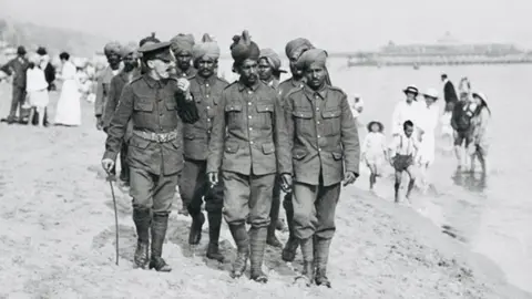 Getty Images A group of Indian soldiers in uniform walking across beach with pier in background and people padding in the sea