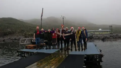 BAS People are standing on a jetty above water. Many are wearing Santa hats and they are holding a "Merry Christmas" banner