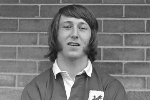 Getty Images Black and white photo of JPR Williams as a young man, wearing a Wales rugby shirt, standing in front of a brick wall. His hair is shoulder-length and he has large sideburns.