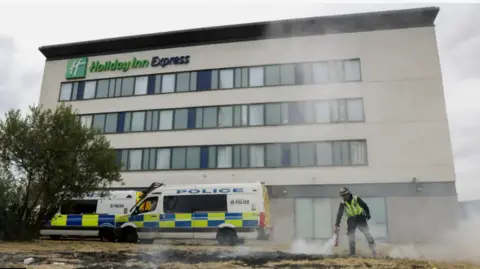 PA Media The Holiday Inn at Manvers, Rotherham,  with two police vans parked in front of it. A man in a high vis jackets is using a fire extinguisher on scorched grass in front of the building.