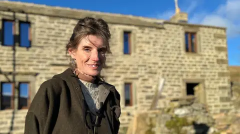 BBC/JULIA LEWIS Amanda Owen has dark hair tied back and wears a green tweed jacket as she stands in front of a stone farmhouse.