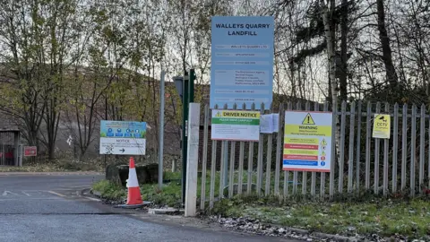 LDRS The entrance to Walleys Quarry - a metal fence on the side of a road has a blue sign attached to it that says "Walleys Quarry Landfill" along with other warning signs. A landfill is barely visible in the background behind a row of trees.