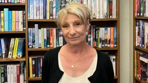 BBC A smiling Jennie Storey looks directly at the camera as she stands in front of a bookshelf full of titles. She is wearing a black cardigan and a white shirt underneath it. She is also wearing a silver necklace with a pendant. 