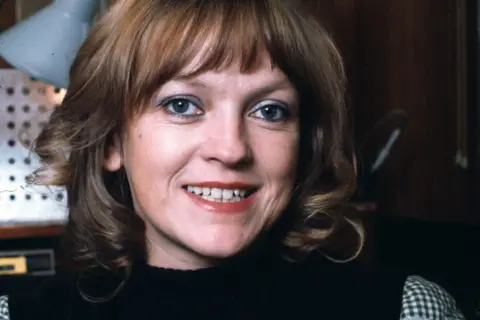 Annie Nightingale photographed in 1976 when she was 36. She has light shoulder-length hair, and is wearing a black tank top over a gingham shirt. She is in a studio and is looking at the camera with a somewhat fixed grin.