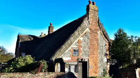 Getty Images A thatched roof building with red brick and grey stones. The cottage has two small windows and a wooden door.