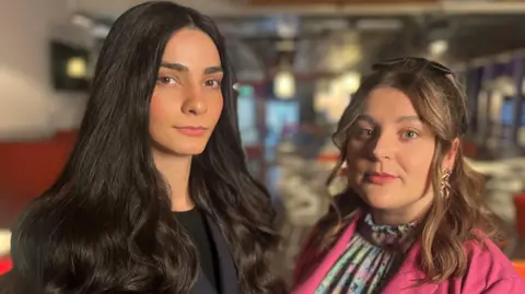 BBC Ellie Wilson & Hannah McLaughlan are photographed looking directly at the camera. The background is blurred out but it is the BBC Scotland newsroom. Ellie has long dark hair and is wearing a dark suit. Ellie is smaller, with shoulder-length hair and a side-parting. She is wearing a pink jacket and has a colourful shirt.