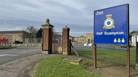 PA Media A blue RAF Scampton sign stands at the entrance to the site with an RAF logo and the badge of RAF Scampton. There are two brick gate posts at the entrance and buildings in the background.