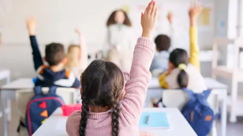Getty Images A shot of primary school-aged children from the back raising their hands as they are asked a question by a teacher