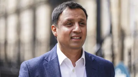 PA Media Anas Sarwar, with dark hair and wearing a blue jacket and white shite, walks along a street